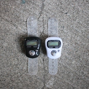 May include: Two digital tally counters, one black and one white, each with a clear wrist strap. The counters display "TALLY COUNTER" above a digital screen with "COUNT" and "RESET" buttons. The background is a textured gray surface.