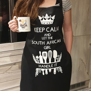 May include: Black apron with white text that reads "Keep Calm and Let the South African Girl Handle It". The apron features a crown, kitchen utensils, and floral designs.