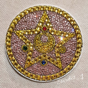 May include: A pink and gold star with a crescent moon and four gems in the centre. The star is surrounded by a gold circle with small rhinestones.