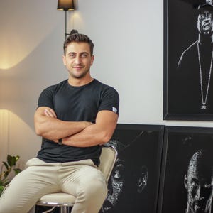 May include: A man wearing a black t-shirt and beige pants sits in front of a wall with three framed black and white portraits. The portraits are of people wearing hats and have a white outline.