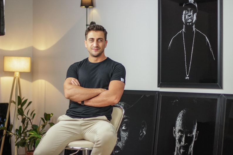 May include: A man wearing a black t-shirt and beige pants sits in front of a wall with three framed black and white portraits. The portraits are of people wearing hats and have a white outline.