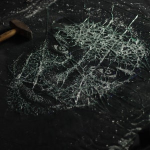 May include: A close-up of a black surface with a shattered glass pane. The glass is broken into many small pieces, creating a spiderweb pattern. A hammer lies on the surface to the left of the broken glass.