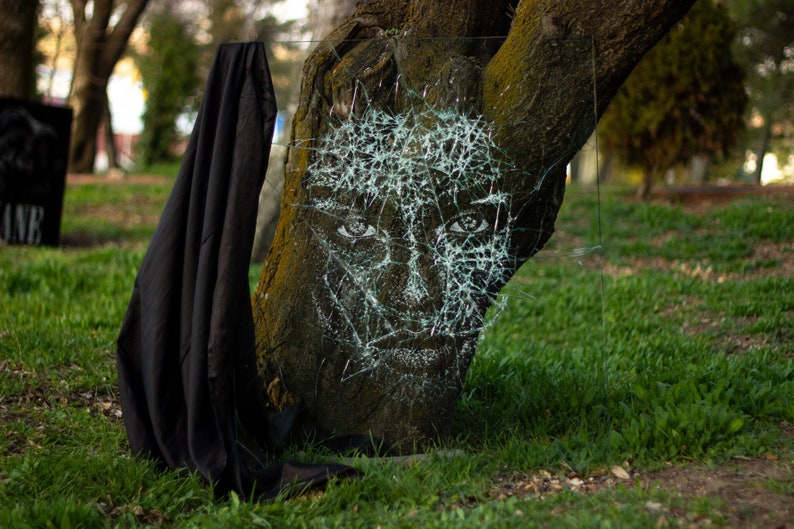 May include: A black cloth drapes over a tree trunk with a face drawn on it in white paint. The face is covered in a web-like pattern of cracks, creating a unique and artistic effect.