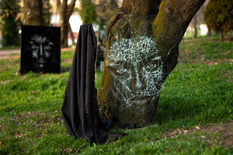 May include: A white face drawn on a tree trunk with broken glass shards covering the face. The glass is arranged in a web-like pattern. A black cloth is draped over the tree trunk.