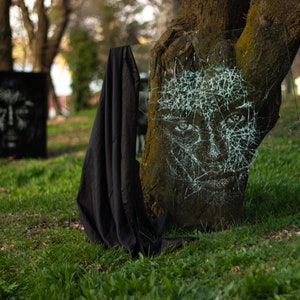 May include: A tree trunk with a face drawn on it with white paint. The face is covered in a web-like pattern of cracks. A black cloth is draped over the base of the tree.
