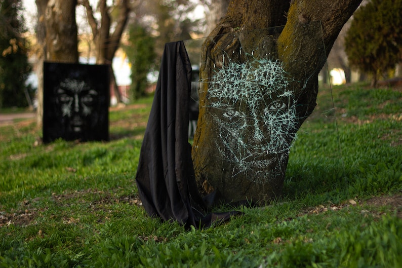 May include: A tree trunk with a face drawn on it with white paint. The face is covered in a web-like pattern of cracks. A black cloth is draped over the base of the tree.