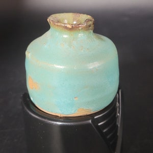 May include: A small, teal-colored ceramic vase with a brown rim, sitting on a black stand.
