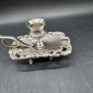 May include: A silver-colored candlestick holder with an ornate, rectangular base and a handle. The candle cup is circular, and the base features decorative scrollwork and small feet. The overall design is vintage and elegant.