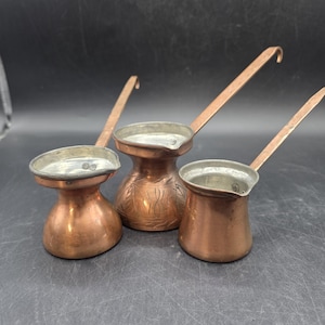 May include: Three copper coffee pots with long handles. The pots have a small, round base and a wider top. The middle pot has an embossed design. The handles are curved at the end.