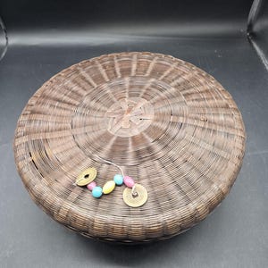 May include: Round, woven basket with a lid. The basket is brown and has a circular design. A decorative string with colorful beads and two gold-colored coins is attached to the side.
