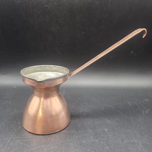 May include: A copper-colored Turkish coffee pot with a long handle. The pot has a rounded base that tapers to a narrow neck and a wide, open top. The handle is attached to the side and curves upward at the end.