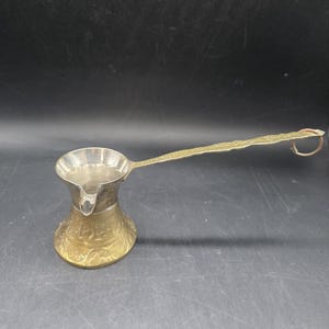 May include: A silver and brass coffee pot with a long, textured handle. The pot has a wide base and a narrow neck, with a spout for pouring. The handle has a loop at the end. The pot is likely used for making Turkish coffee.
