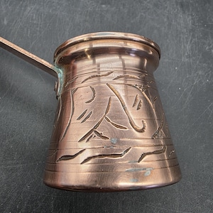 May include: A copper-colored Turkish coffee pot with a long handle. The pot features etched floral designs around the body. The pot is made of copper and has a shiny finish. The handle is attached to the side of the pot.