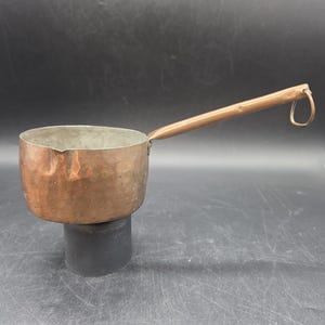 May include: A copper saucepan with a long, wooden handle and a small loop at the end. The pan has a rounded base and a slightly flared rim. The copper has a warm, reddish-brown color, and the handle is a lighter brown.