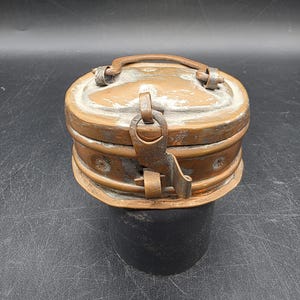 May include: An antique copper-toned metal box with an oval shape and a hinged lid. The box features a handle on top and a clasp closure. The metal shows signs of age and wear, with some areas of discoloration. The box is sitting on a black surface.