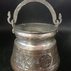 May include: An antique metal cauldron with a curved handle and decorative floral patterns. The cauldron has a silver-colored interior and a bronze-colored exterior with etched designs. The handle is attached with ornate, curved brackets.