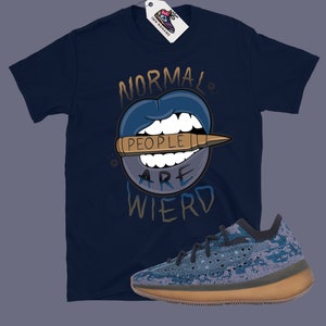 Op de afbeelding: Een donkerblauw t-shirt met een afbeelding van een mond met een kogel waarop staat "Mensen zijn raar". Een paar blauwe en bruine sneakers bevinden zich in de rechterbenedenhoek van de afbeelding.