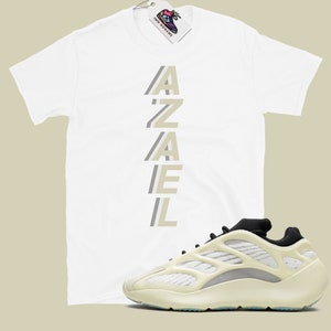 May include: White t-shirt with the word "AZAEL" printed vertically in a light beige color. A pair of white and beige sneakers with black accents.