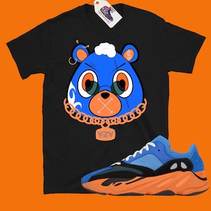 May include: Black t-shirt with a blue cartoon bear wearing a gold chain and a YZY pendant. The bear has a dollar sign on its ear and a stitched-up mouth. A pair of orange and blue Yeezy sneakers are in the bottom right corner of the image.