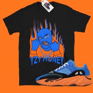 May include: Black t-shirt with a blue masked figure with orange flames and the text "YZY MONEY".  A pair of blue and orange sneakers are also pictured.
