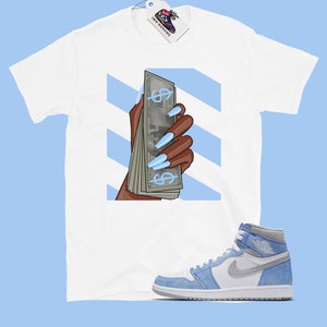 May include: White t-shirt featuring a graphic of a hand holding a stack of cash with blue dollar signs and long blue nails. The background is blue and white stripes. A pair of light blue and white trainers are also pictured.