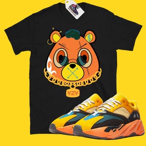May include: Black t-shirt featuring an orange bear graphic with gold chain, glasses, and dollar sign. Paired with orange, yellow, and blue sneakers. The tag reads "IMN NONAME".