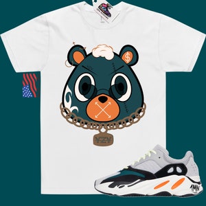 May include: White t-shirt featuring a cartoon illustration of a green bear wearing a gold chain with a YZY pendant. The bear has a dollar sign on its ear and an X over its mouth. The t-shirt is paired with grey, green, and orange trainers.