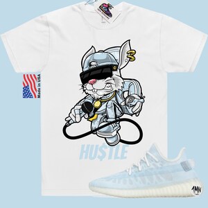 May include: White t-shirt featuring a cartoon rabbit wearing sunglasses and a backwards hat holding a microphone. The text "HUSTLE" is printed below the rabbit. A pair of light blue and white trainers are shown below the t-shirt.
