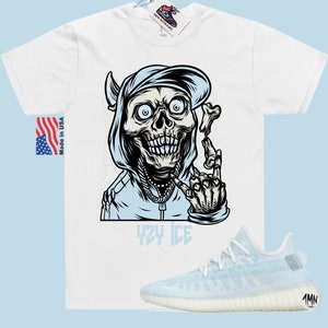 May include: White t-shirt with a graphic of a cartoon skeleton wearing a blue hoodie and a baseball cap. The skeleton is holding a bone in its hand and has the words "YZY ICE" printed below it. A pair of light blue and white sneakers are also pictured.