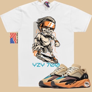 May include: White t-shirt with a cartoon illustration of a person wearing a virtual reality headset and riding a skateboard. The text "YZY 700" is printed below the illustration. A pair of tan, orange, and blue sneakers are shown below the t-shirt.