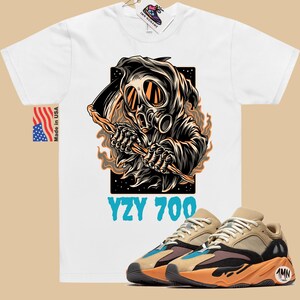 May include: White t-shirt with a graphic of a hooded figure wearing a gas mask and holding a scythe. The text "YZY 700" is printed below the graphic. A pair of tan and orange sneakers with black accents are shown below the t-shirt.