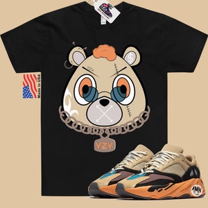 May include: Black t-shirt featuring a cartoon bear wearing a chain and a tag that reads "YZY". The bear has a stitched-up face with a dollar sign on its ear. A pair of tan, orange, and blue trainers with a black sole and a tag that says "1MN" on the side.