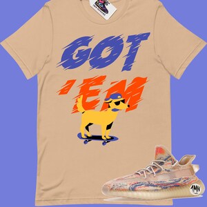 May include: A tan t-shirt with the text "GOT 'EM" in blue and orange. A cartoon dog wearing sunglasses and a blue hat is skateboarding. A pair of beige and multi-coloured trainers are shown below the t-shirt.
