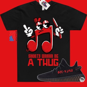 Puede incluir: Camiseta negra con un personaje de nota musical roja sosteniendo un cuchillo y el texto "Shorty Wanna Be A Thug". Debajo de la camiseta se muestra un par de zapatillas Yeezy Boost 350 V2 negras con detalles rojos.