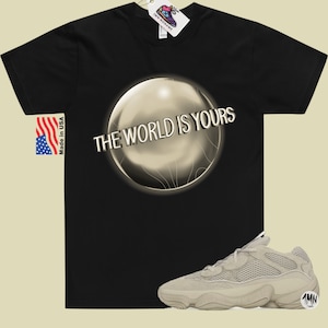 May include: Black t-shirt with a silver orb graphic and the text "THE WORLD IS YOURS". A pair of beige sneakers with a mesh upper and a thick sole.