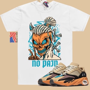 May include: White t-shirt with a graphic of a skull with dreadlocks and the text "no pain". The skull is wearing a blue bandana and has a blue and orange color scheme. A pair of tan, orange, and black sneakers are shown below the t-shirt.