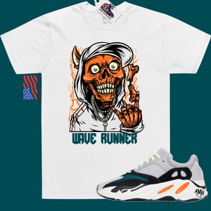 Puede incluir: Camiseta blanca con un gr&aacute;fico de una calavera con una sudadera con capucha y una gorra puesta hacia atr&aacute;s. La calavera es naranja y tiene llamas a su alrededor. El texto "WAVE RUNNER" est&aacute; impreso debajo del gr&aacute;fico. Un par de zapatillas grises, verdes y naranjas se muestran debajo de la camiseta.