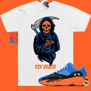 May include: White t-shirt featuring a Grim Reaper graphic holding a blue rose, with the text "YZY DEATH". A pair of blue, black, and orange trainers are in the foreground. The shirt has a tag that says "1MN SONAME".