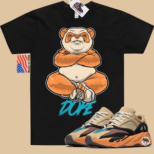 Puede incluir: Camiseta negra con un panda de dibujos animados que lleva un sombrero y est&aacute; sentado con las piernas cruzadas. La camiseta tiene la palabra "Dope" escrita en azul. Un par de zapatillas de deporte de color beige, naranja y azul con la palabra "1MN" en el lateral se muestran debajo de la camiseta.