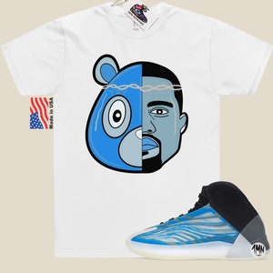 May include: White t-shirt with a blue and grey cartoon image of a bear and a man's face. The image has a crown of thorns on top.  A pair of blue and white sneakers with black accents and a white sole.