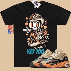 Puede incluir: Camiseta negra con un esqueleto de dibujos animados que lleva un sombrero y patina en monopat&iacute;n. El texto "YZY 100" est&aacute; impreso debajo de la imagen. Un par de zapatillas de deporte de color beige, naranja y azul se muestran debajo de la camiseta.