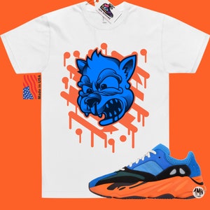 May include: White t-shirt with a blue cartoon wolf graphic and orange paint drips. The t-shirt is paired with orange and blue sneakers with a black swoosh.