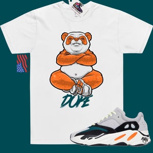 Pu&ograve; includere: Maglietta bianca con un panda dei cartoni animati che indossa pantaloni arancioni e siede con le braccia incrociate. Il testo "DUPE" &egrave; scritto in verde sotto il panda. Un paio di scarpe da ginnastica grigie, nere, arancioni e verdi sono mostrate sotto la maglietta.
