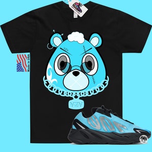 May include: Black t-shirt with a blue cartoon bear wearing a chain with a YZY pendant.  The bear has a stitched-up face and a cloud on its head.  A pair of blue and black YZY sneakers are below the t-shirt.