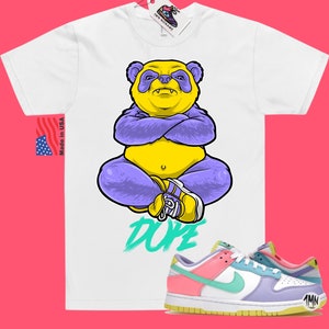 May include: White t-shirt with a cartoon illustration of a yellow and purple panda with the word "DOPE" printed below. A pair of pink, green, and purple Nike low-top sneakers with a yellow sole.