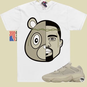 May include: White t-shirt with a graphic of a bear and Kanye West's face. The graphic is split down the middle, with the bear on the left and Kanye on the right. The bear is wearing a crown of thorns.  A pair of beige sneakers with a mesh upper and a thick sole are shown below the t-shirt.