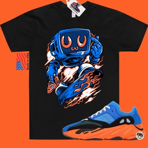 May include: Black t-shirt with a cartoon illustration of a blue robot holding a video game controller and riding a flaming skateboard. The robot has a blue television screen for a head with a cartoon face. The t-shirt is paired with a pair of orange and blue sneakers with the text "1MN" on the side.