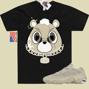 May include: Black t-shirt with a cartoon bear wearing a gold chain and a pendant that says "Yeezy". The bear has a white patch on its head and a stitched-up face. The t-shirt is paired with beige sneakers with a mesh design.