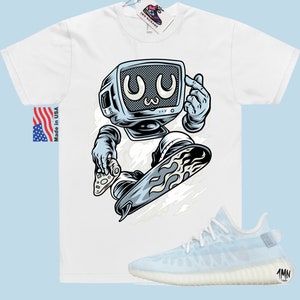 May include: White t-shirt with a cartoon illustration of a robot with a television for a head, riding a snowboard. The robot is wearing a blue and white spacesuit and has a blue and white snowboard with flames on the bottom. The t-shirt has the text "Made in USA" on the left side. The image also includes a pair of white and light blue sneakers with the text "1MN" on the side.