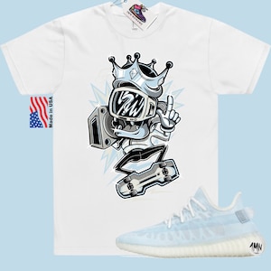 Puede incluir: Camiseta blanca con una ilustraci&oacute;n de dibujos animados de un personaje que lleva una corona y sostiene un boombox. El personaje est&aacute; haciendo skateboard. La camiseta se combina con un par de zapatillas de deporte azul claro con suela blanca.